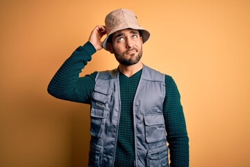 Handsome tourist man with beard on vacation wearing explorer hat over yellow background confuse and wondering about question. Uncertain with doubt, thinking with hand on head. Pensive concept.