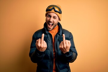 Young handsome skier man with beard wearing snow sportswear and ski goggles Showing middle finger...
