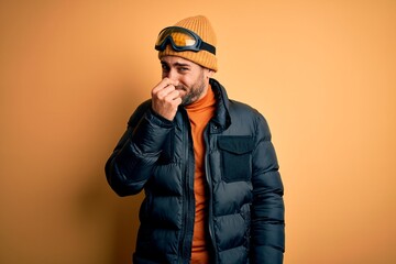 Young handsome skier man with beard wearing snow sportswear and ski goggles smelling something stinky and disgusting, intolerable smell, holding breath with fingers on nose. Bad smell