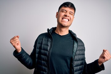 Young handsome hispanic man wearing winter coat standing over white isolated background very happy and excited doing winner gesture with arms raised, smiling and screaming for success. Celebration.