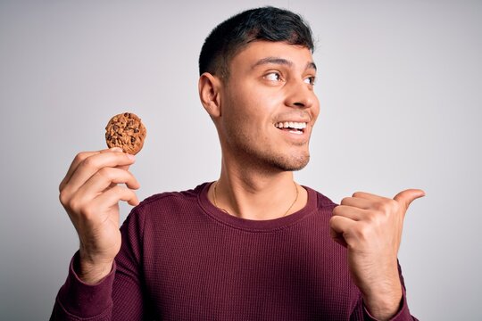 Young hispanic man eating chocolate chips cookie over isolated background pointing and showing with thumb up to the side with happy face smiling