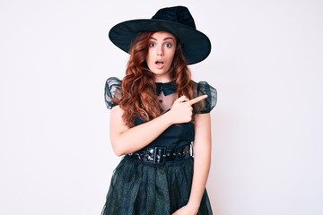 Young beautiful woman wearing witch halloween costume surprised pointing with finger to the side, open mouth amazed expression.