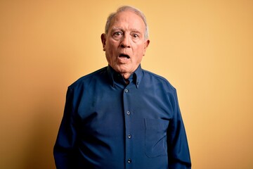 Grey haired senior man wearing casual blue shirt standing over yellow background In shock face, looking skeptical and sarcastic, surprised with open mouth