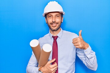 Young handsome man wearing architect hardhat holding blueprint smiling happy and positive, thumb up doing excellent and approval sign