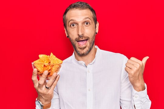 Young handsome man holding nachos potato chips pointing thumb up to the side smiling happy with open mouth