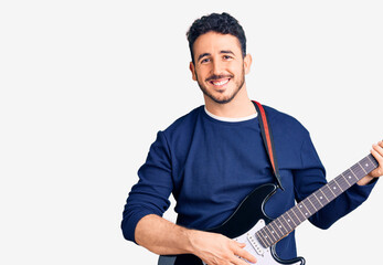 Young hispanic man playing electric guitar looking positive and happy standing and smiling with a confident smile showing teeth