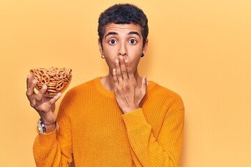Young african amercian man holding pretzels covering mouth with hand, shocked and afraid for...