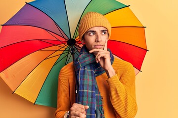 Young african amercian man holding colorful umbrella serious face thinking about question with hand on chin, thoughtful about confusing idea