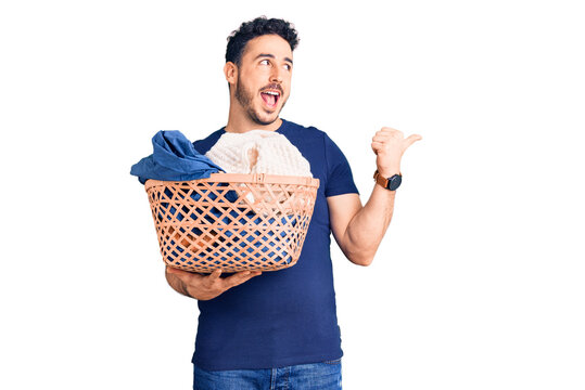 Young hispanic man holding laundry basket pointing thumb up to the side smiling happy with open mouth