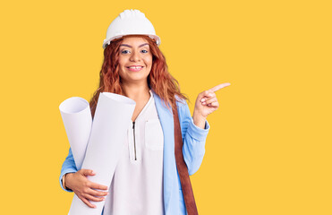 Young latin woman wearing architect hardhat and leather bag holding blueprints smiling happy pointing with hand and finger to the side