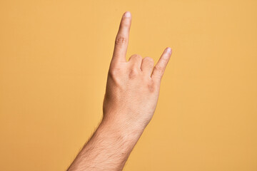 Hand of caucasian young man showing fingers over isolated yellow background gesturing rock and roll symbol, showing obscene horns gesture