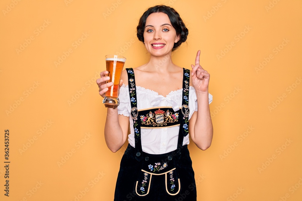 Wall mural Young beautiful german woman with blue eyes wearing octoberfest dress drinking glass of beer surprised with an idea or question pointing finger with happy face, number one