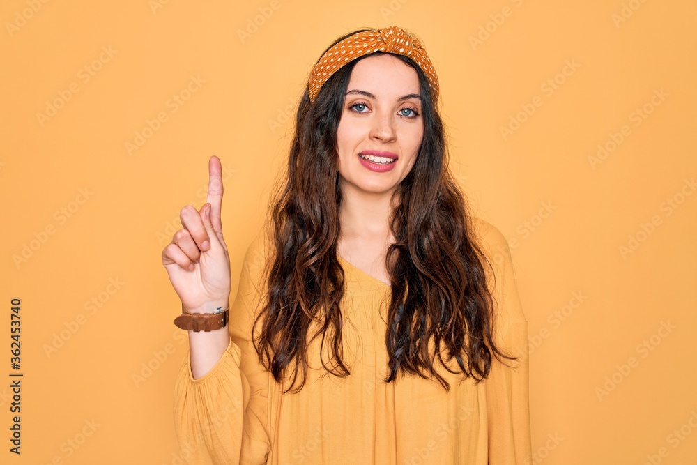 Wall mural Young beautiful woman wearing casual t-shirt and diadem standing over yellow background showing and pointing up with finger number one while smiling confident and happy.