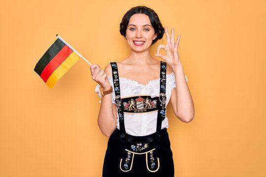 Young patriotic german woman with blue eyes wearing octoberfest dress holding germany flag doing ok sign with fingers, excellent symbol