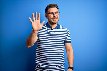 Naklejka premium Young man with blue eyes wearing glasses and casual striped t-shirt over blue background showing and pointing up with fingers number five while smiling confident and happy.