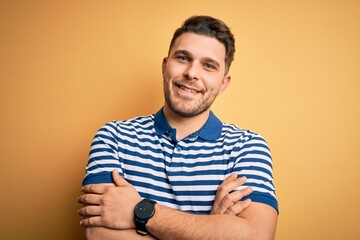 Young man with blue eyes wearing casual striped t-shirt over yellow background happy face smiling with crossed arms looking at the camera. Positive person.
