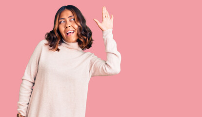 Young beautiful mixed race woman wearing winter turtleneck sweater waiving saying hello happy and smiling, friendly welcome gesture © Krakenimages.com