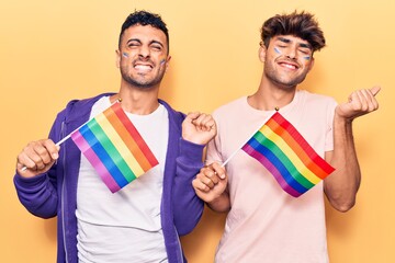 Young gay couple holding rainbow lgbtq flags screaming proud, celebrating victory and success very excited with raised arm