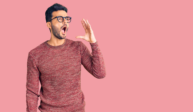 Young handsome hispanic man wearing winter sweater and glasses shouting and screaming loud to side with hand on mouth. communication concept.