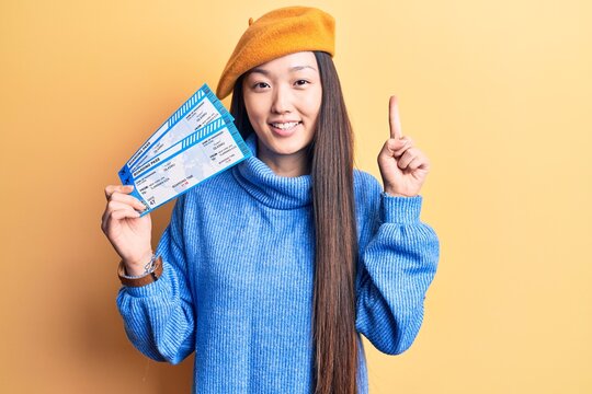 Young beautiful blonde woman holding airline boarding pass smiling with an idea or question pointing finger with happy face, number one