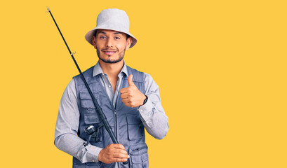 Handsome latin american young man wearing fisherman equipment smiling happy and positive, thumb up doing excellent and approval sign