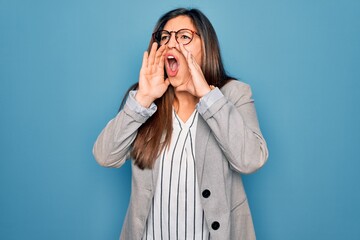 Young hispanic business woman wearing glasses standing over blue isolated background Shouting angry out loud with hands over mouth