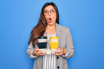 Young hispanic intern business woman holding coffee over blue background scared in shock with a...