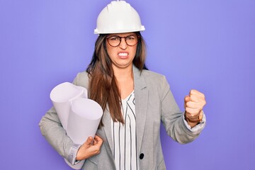 Young hispanic architect woman wearing builder hat and holding blueprints over yellow background annoyed and frustrated shouting with anger, crazy and yelling with raised hand, anger concept