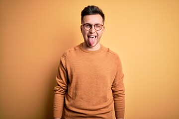 Young handsome caucasian man wearing glasses and casual winter sweater over yellow background sticking tongue out happy with funny expression. Emotion concept.
