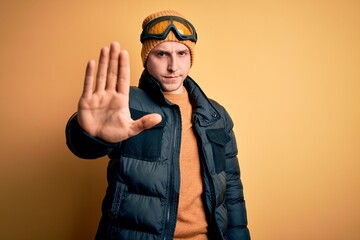 Young handsome caucasian man wearing hat, coat and ski glasses for winter and snow weather doing stop sing with palm of the hand. Warning expression with negative and serious gesture on the face.