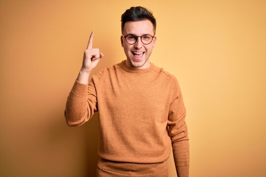 Young handsome caucasian man wearing glasses and casual winter sweater over yellow background pointing finger up with successful idea. Exited and happy. Number one.