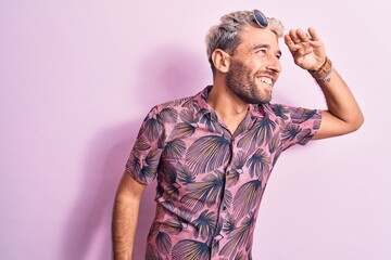 Handsome blond man on vacation wearing casual shirt and sunglasses over pink background very happy and smiling looking far away with hand over head. Searching concept.