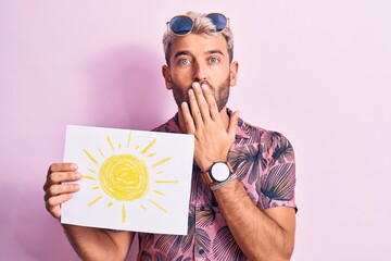 Handsome blond tourist man on vacation wearing sunglasses holding paper with sun draw covering...