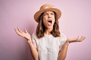 Young beautiful blonde girl wearing summer hat over pink isolated background crazy and mad shouting and yelling with aggressive expression and arms raised. Frustration concept.