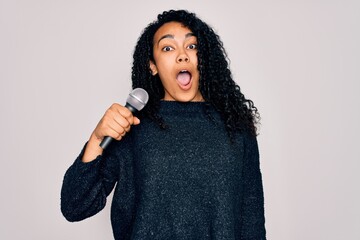 Young african american curly singer woman singing using microphone over white background scared in...
