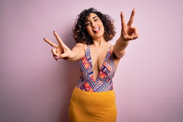 Young beautiful arab woman on vacation wearing swimsuit and sunglasses over pink background smiling with tongue out showing fingers of both hands doing victory sign. Number two.