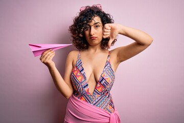 Young beautiful curly arab woman on vacation wearing swimsuit holding pink paper airplane with angry face, negative sign showing dislike with thumbs down, rejection concept