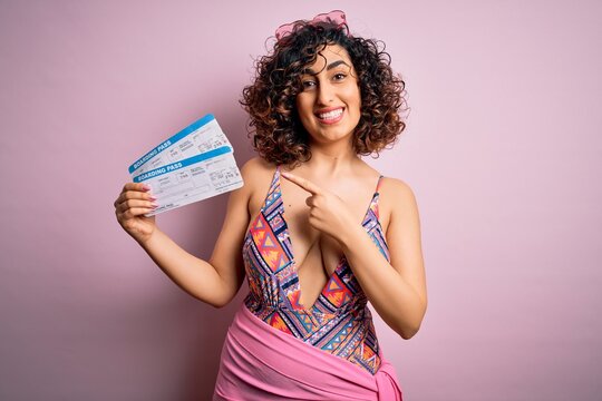 Young beautiful tourist arab woman on vacation wearing swimsuit holding boarding pass airline very happy pointing with hand and finger