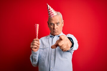 Middle age man wearing funny hat using trumpet celebrating birthday over red background pointing with finger to the camera and to you, hand sign, positive and confident gesture from the front