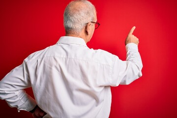 Middle age handsome hoary man wearing casual shirt and glasses over red background Posing backwards pointing ahead with finger hand