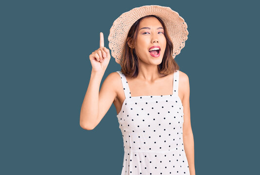 Young beautiful chinese girl wearing summer hat pointing finger up with successful idea. exited and happy. number one.