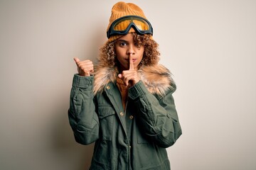 Young african american skier woman with curly hair wearing snow sportswear and ski goggles asking to be quiet with finger on lips pointing with hand to the side. Silence and secret concept.