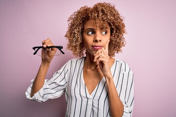 African american optical woman with curly hair holding glasses over isolated pink background...