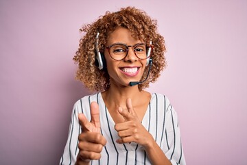 African american curly call center agent woman working using headset over pink background pointing...