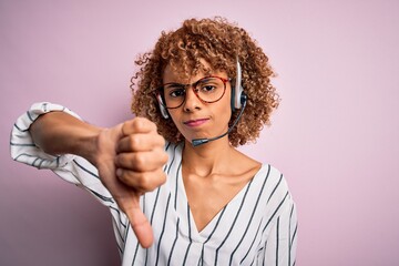 African american curly call center agent woman working using headset over pink background looking...