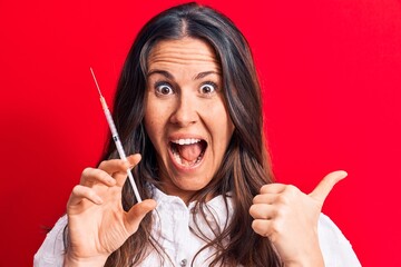 Young beautiful brunette woman holding syringe  over isolated red background pointing thumb up to...