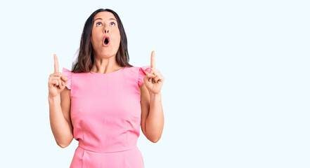 Young beautiful brunette woman wearing casual dress amazed and surprised looking up and pointing with fingers and raised arms.