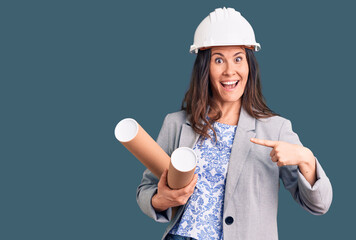 Young beautiful brunette woman wearing architect hardhat holding blueprint smiling happy pointing with hand and finger