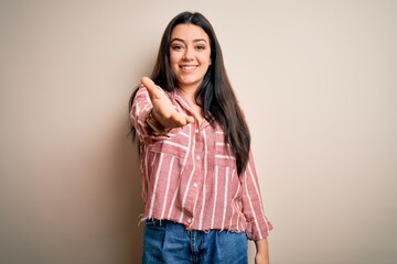 Young brunette woman wearing casual striped shirt over isolated background smiling friendly offering handshake as greeting and welcoming. Successful business.