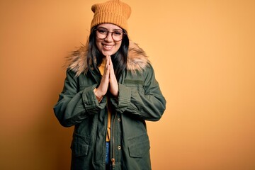 Young brunette woman wearing glasses and winter coat with hat over yellow isolated background praying with hands together asking for forgiveness smiling confident.